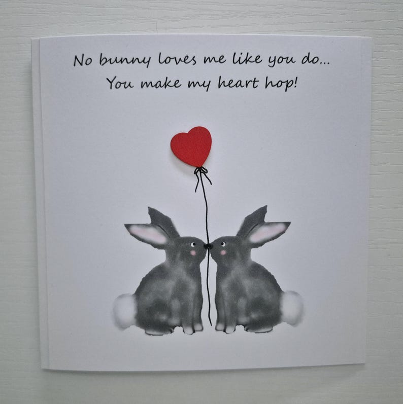 Może przedstawiać: Kartka z życzeniami z czerwonym balonem w kształcie serca i dwoma szarymi kr&oacute;likami, kt&oacute;re się całują. Na kartce napisano "No bunny loves me like you do... You make my heart hop!"