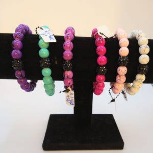 May include: Six colorful beaded bracelets with black accents and a rhinestone charm. The bracelets are displayed on a black stand. The price tags are visible with the text "$11.95" and "$11.95".