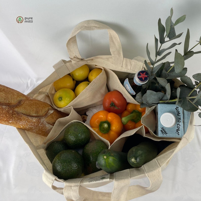 May include: A beige canvas tote bag with multiple compartments filled with groceries. The bag contains lemons, avocados, a baguette, bell peppers, a tomato, a bottle of juice, and a carton of milk. The bag has the words "pure mitti" printed on the front.