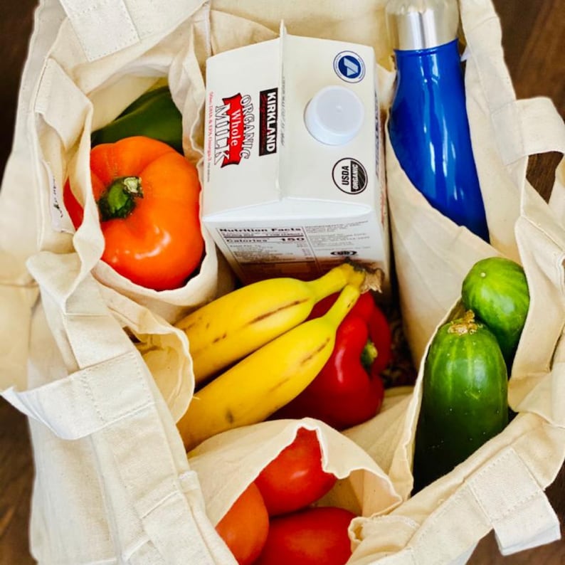May include: A white reusable tote bag filled with groceries, including a carton of Kirkland Signature Organic Whole Milk, bananas, tomatoes, peppers, and cucumbers.