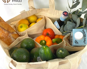 May include: A beige canvas tote bag with internal compartments filled with groceries including a baguette, lemons, avocados, a cucumber, bell peppers, a tomato, a glass bottle, a carton labeled wow no cow, and green eucalyptus branches.