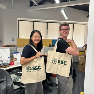 May include: Two people are holding reusable shopping bags with the logo "SSC Sustainability Service Corps" printed on them. The bags are beige with green text and a green tree logo.