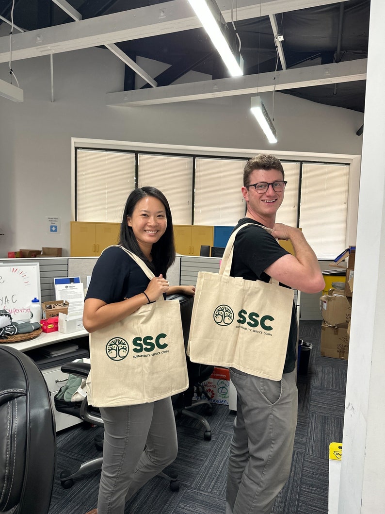 May include: Two people are holding reusable shopping bags with the logo "SSC Sustainability Service Corps" printed on them. The bags are beige with green text and a green tree logo.