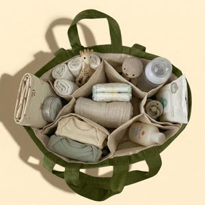 May include: An olive green tote bag filled with baby essentials. The bag contains diapers, baby clothes, a bottle, wipes, a pacifier, a giraffe toy, and a crocheted doll. The bag has multiple compartments for organization.