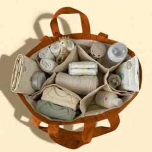 May include: A burnt orange tote bag with multiple compartments, filled with baby essentials. Items include diapers, baby clothes, a bottle, wipes, a pacifier, a giraffe toy, and a small stuffed animal. The bag has two handles.