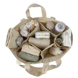 May include: A beige canvas tote bag with multiple compartments, filled with baby essentials. Items include diapers, baby clothes, a giraffe toy, a pacifier, wipes, a bottle, and a small jar of cream. The bag has two handles.