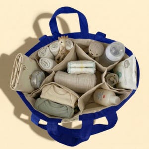 May include: A navy blue tote bag with multiple compartments, filled with baby essentials. Items include diapers, baby clothes, a bottle, wipes, a pacifier, and a giraffe toy. The bag has two handles and a light beige interior.