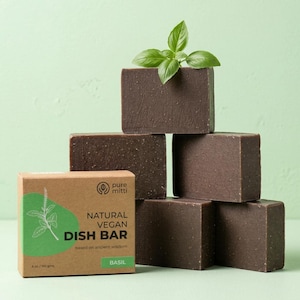 May include: A stack of five rectangular dark brown dish bars with a textured surface, topped with a fresh green basil sprig. A brown cardboard box sits in front, labeled Pure Mitti Natural Vegan Dish Bar Basil, 6 oz / 160 gms.