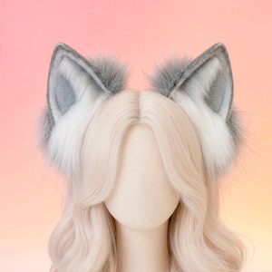 May include: A mannequin head with long, wavy, light blonde hair, adorned with a pair of gray and white faux fur wolf ears. The ears are detailed with a fluffy texture and sit atop the head. The background is a gradient of pink and yellow.