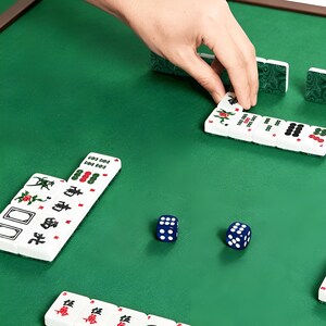 May include: A mahjong set featuring white tiles with green, red, and black character and symbol engravings, arranged on a green felt surface alongside two blue six-sided dice with white pips, showing a game in progress with various tile patterns.