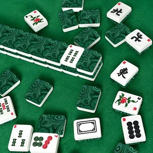 May include: A set of mahjong tiles featuring a deep green malachite-style marbled pattern on the backs. The front faces are white with black, red, and green engraved characters, numbers, and floral motifs, arranged in a scattered layout.