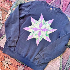 May include: A dark gray sweatshirt featuring a large, star-shaped appliqué. The star is composed of various fabric patches in shades of pink, cream, and green. The sweatshirt has long sleeves and a crew neck. The overall style is casual and handmade.