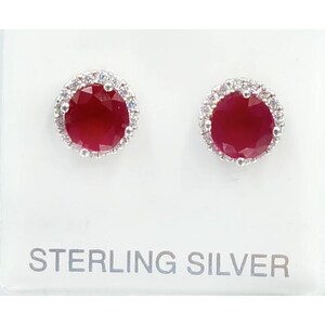 May include: A pair of sterling silver stud earrings. Each earring features a round, deep red gemstone surrounded by a halo of smaller, clear stones. The earrings are displayed on a white surface with the text "STERLING SILVER".