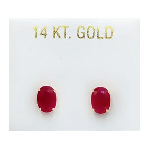 May include: A pair of oval-shaped red gemstone stud earrings set in gold prongs, mounted on a white display card with the text 14 KT. GOLD printed in gold lettering at the top. The earrings feature faceted red stones with a polished finish.