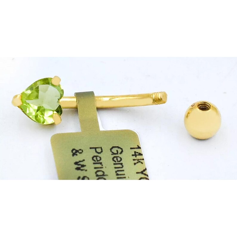 May include: A 14k yellow gold belly button ring with a green peridot heart-shaped gemstone. The ring is attached to a small gold ball.