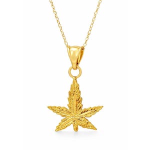 May include: A gold-colored necklace featuring a detailed cannabis leaf pendant. The pendant is suspended from a delicate chain. The leaf design has intricate veining and a textured surface, reflecting light. The necklace is likely made of gold or a gold-toned metal.