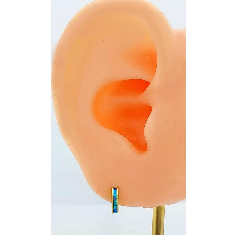 May include: A gold-coloured rectangular earring with a blue and green opal inlay is shown on an ear. The earring is a small, vertical bar shape. The ear is a light peach colour, and the background is white.
