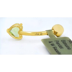 May include: A gold belly button ring with a heart-shaped green gemstone. The ring is marked "14K D" and comes with a tag that reads "Genuine Peridot & W. Sand."