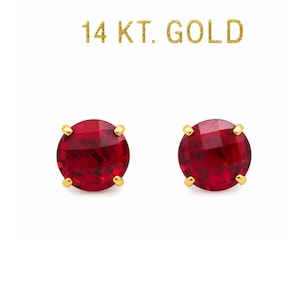 May include: A pair of round, faceted red gemstone stud earrings set in 14 kt gold prongs. The text 14 KT. GOLD is printed in gold lettering above the earrings. Each earring features a circular red stone with a multi-faceted surface.
