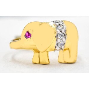 Peut inclure: Bague dorée en forme d'éléphant, ornée d'un œil en rubis et d'une bande de pierres claires. Le bijou est présenté sur fond blanc, mettant en valeur les détails et le design.