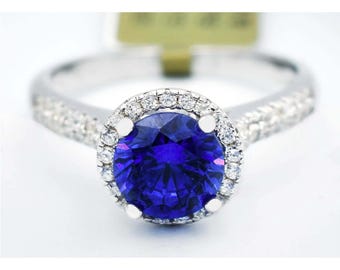 Op de afbeelding: Een zilveren ring met een grote, ronde, diepblauwe edelsteen in het midden, omringd door een halo van kleinere, heldere edelstenen. De ringband is ook bezet met kleine, heldere edelstenen.