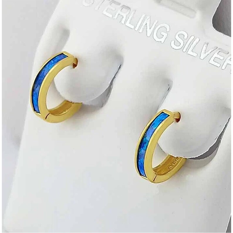May include: A pair of gold-coloured hoop earrings featuring a bright blue inlay. The earrings are presented on a white surface, highlighting their polished finish and the contrast between the gold and blue tones. The earrings are small and delicate.