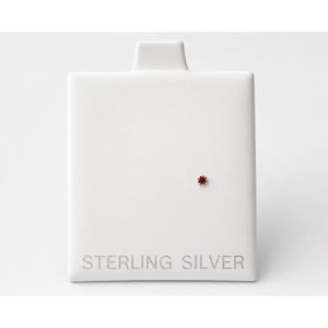 May include: A single small stud earring featuring a tiny, round, deep red gemstone set in a metal prong setting. The earring is mounted on a rectangular white display card with the words STERLING SILVER embossed in gray at the bottom.