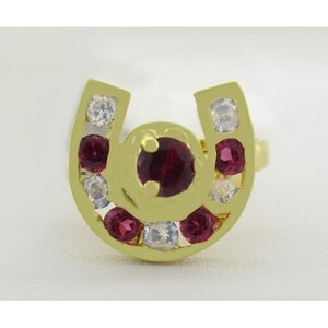 May include: A gold horseshoe ring with red and white gemstones. The ring is set with a large red gemstone in the center and smaller red and white gemstones around the horseshoe.