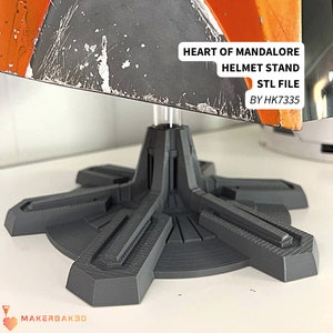 Könnte beinhalten: Ein 3D-gedruckter Ständer für einen Helm aus dunkelgrauem Kunststoff. Der Ständer hat eine runde Basis mit fünf Armen, die nach außen ragen. Der Ständer ist so konzipiert, dass er einen Helm an Ort und Stelle hält. Der Text "HEART OF MANDALORE HELMET STAND STL FILE BY HK7335" ist auf den Ständer gedruckt.