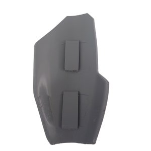 Pu&ograve; includere: Un oggetto di plastica grigio curvo con due cinghie rettangolari attaccate. L'oggetto ha una superficie leggermente strutturata e sembra essere un componente protettivo o parte di un elemento pi&ugrave; grande. La forma &egrave; asimmetrica, con una parte superiore appuntita e inferiore arrotondata.