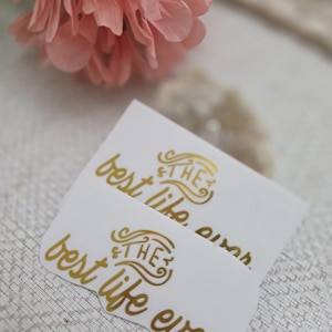 May include: Two white rectangular stickers with rounded corners featuring gold metallic lettering. The text reads "THE best life ever" in a stylized script font with decorative flourishes around the word "THE".