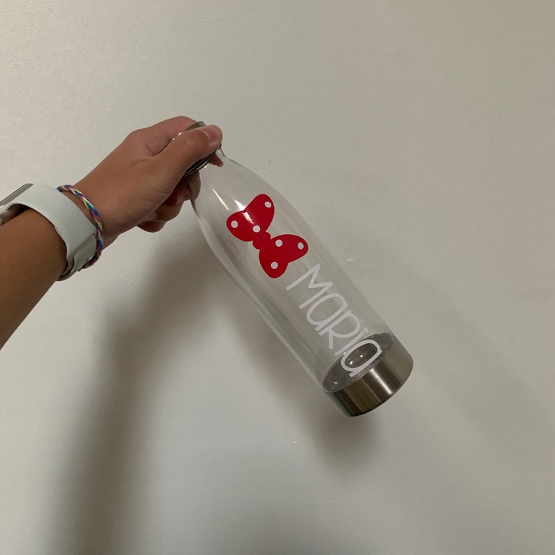 K&ouml;nnte beinhalten: Transparente Wasserflasche aus Plastik mit einem roten Schmetterlingsaufkleber und dem Namen "Maria" in wei&szlig;. Die Flasche hat einen silbernen Deckel.