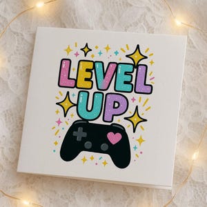 Può includere: Un biglietto bianco con la scritta "LEVEL UP" in lettere a bolle colorate, un controller di gioco nero e stelle. Il biglietto è decorato con simboli di cuore e croce.