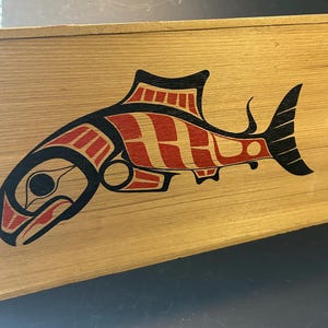 Peut inclure: Une boîte en bois avec un saumon stylisé rouge et noir peint sur le dessus. Le saumon est tourné vers la gauche et a un gros œil.