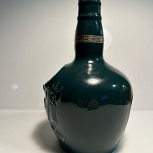 May include: A dark green ceramic bottle with a silver top and a raised design on the side. The bottle is empty and appears to be a vintage decanter.