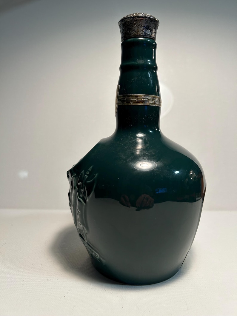 May include: A dark green ceramic bottle with a silver top and a raised design on the side. The bottle is empty and appears to be a vintage decanter.