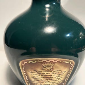 May include: A dark green ceramic bottle with a gold label. The label features a gold crown and a carriage with a wheel. The text on the label reads "By an ancient custom, originating in the Royal Navy, a salute of twenty-one guns is the prerogative of the Reigning Sovereign. Royal Salute Whisky, created to celebrate the Coronation of Her Majesty Queen Elizabeth symbolizes this honour."
