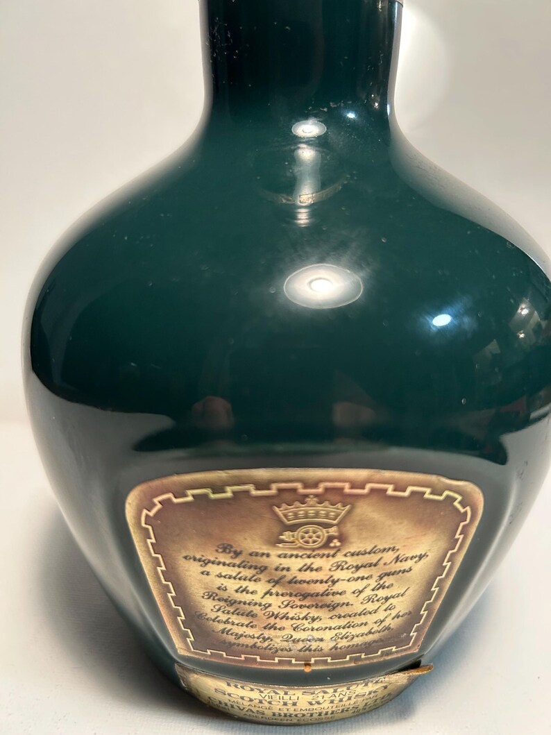 May include: A dark green ceramic bottle with a gold label. The label features a gold crown and a carriage with a wheel. The text on the label reads "By an ancient custom, originating in the Royal Navy, a salute of twenty-one guns is the prerogative of the Reigning Sovereign. Royal Salute Whisky, created to celebrate the Coronation of Her Majesty Queen Elizabeth symbolizes this honour."