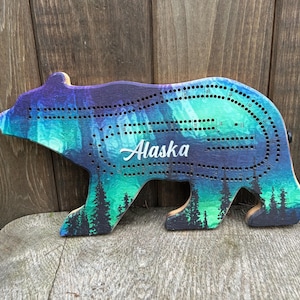 May include: Wooden cribbage board in the shape of a bear, painted with a vibrant scene of the Northern Lights. The design features shades of blue, green, and purple, with the word "Alaska" in white script. The board has holes for pegs.