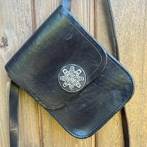 Peut inclure: Sac bandoulière en cuir noir avec un emblème décoratif argenté sur le rabat. Le sac a une longue bandoulière réglable et une forme rectangulaire aux coins arrondis. Le sac est suspendu sur un fond en bois.