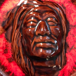 May include: A brown ceramic wall plaque featuring a relief sculpture of a Native American man's face. The background is red with black and white accents.