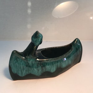 May include: A green glazed ceramic canoe planter with a raised prow. The planter has a dark green glaze with lighter green streaks.