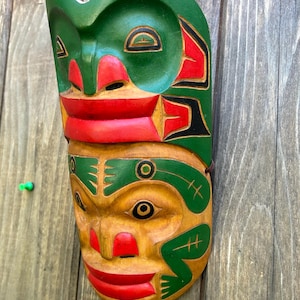 May include: A hand-carved wooden mask with a green top and tan lower section. The mask has red accents on the mouth and nose, with black detailing. It is likely a decorative item or a piece of art, possibly from a cultural tradition.