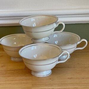 May include: Four white teacups with gold trim, stacked on top of each other. The cups have a classic design and are perfect for a tea party.