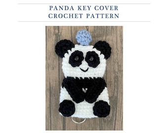 May include: A crocheted panda key cover with black and white fur, black eyes, and a blue pom-pom on its head. The panda is holding a key ring.