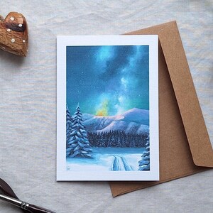 Peut inclure: Une carte de vœux avec une scène hivernale d'une forêt enneigée avec un chemin qui la traverse. Le ciel est bleu foncé avec une aurore boréale jaune et verte brillante.