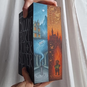 May include: A book with the title "Callie Hart Quicksilver" on the spine. The book's edges feature colorful illustrations of a house, a sword in water, and a figure in flames. The book is held by a person.