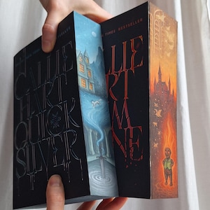 May include: A person holding open a book with a black cover and embossed silver lettering. The book's pages reveal colorful illustrations of a snowy landscape, a sword, and a burning castle. The book's title is "Callie Hart Quicksilver".