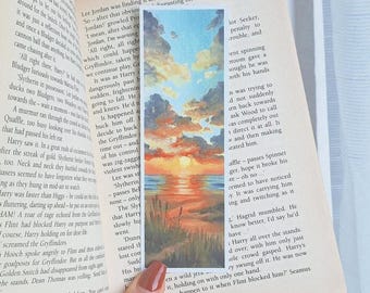 May include: A watercolor bookmark featuring a sunset over a body of water with a sandy beach and green grass. The sky is a vibrant orange and pink with fluffy white clouds. The water is a deep blue with a reflection of the sunset. The beach is a light brown with green grass growing on it.