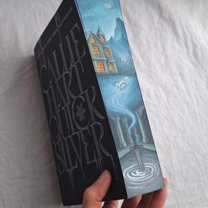 May include: A hardcover book titled "Quicksilver" by Callie Hart. The cover features a detailed illustration of a snowy landscape with a house, a wolf, and a sword. The title is embossed in silver on a black background.